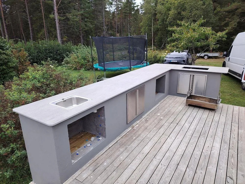 L-formad bänkskiva i matt grå Dekton Keon till utekök på trädäck i Grisslehamn, med rostfri diskho och trädgård bakom.