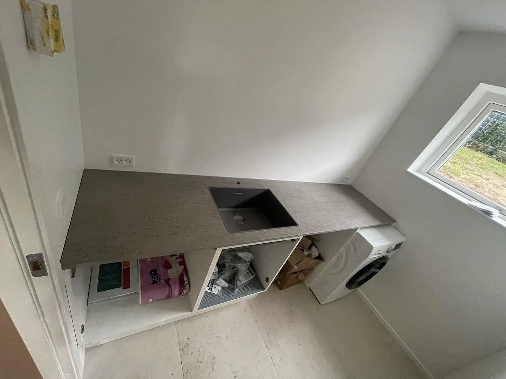 Grå Dekton Keon köksbänkskiva med underlimmad kompositdiskho i ett mindre kök under montering, med fönster och tvättmaskin, i Kr. Hyllinge.