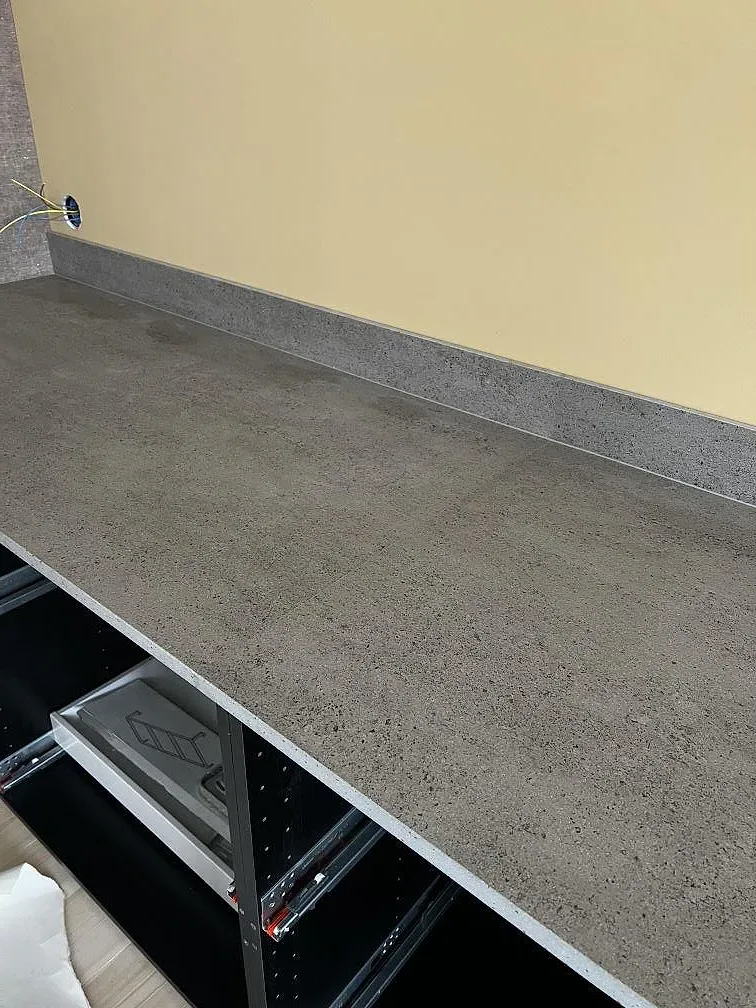 Grå Dekton Keon köksbänkskiva i matt yta längs en gul vägg med öppna lådstommar under, fotograferad under montering i Stora Höga.