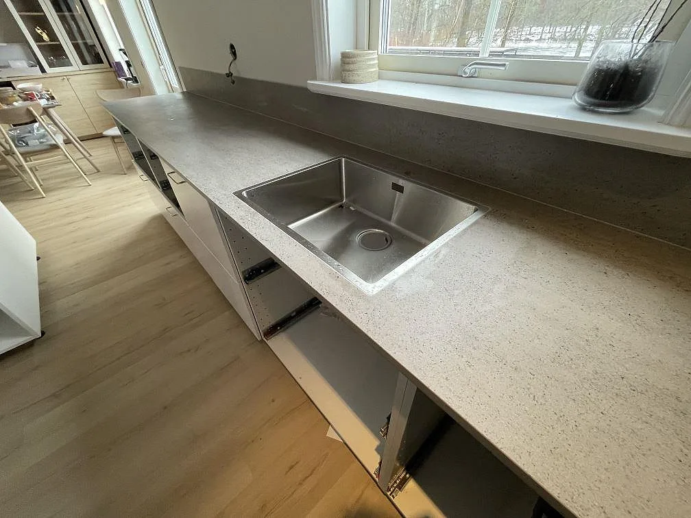 Underlimmad rostfri diskho i grå Dekton Keon-bänkskiva under fönstret i ett kök i Surahammar.