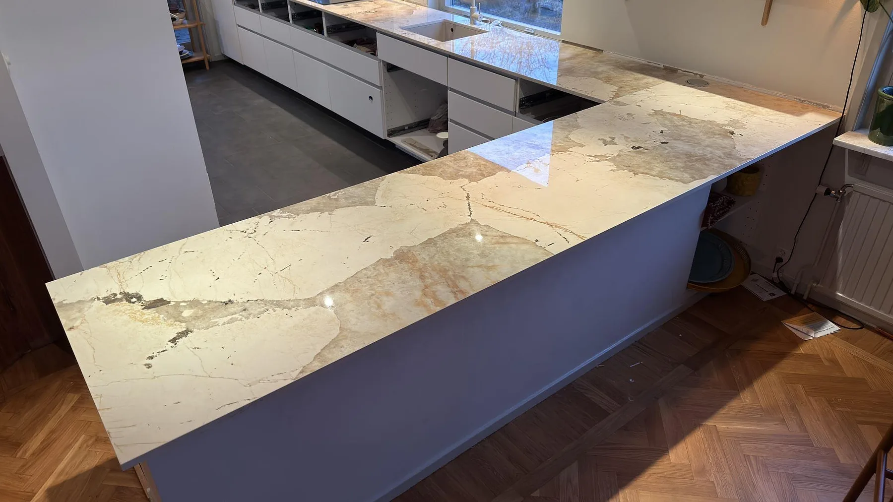 Polerad köksbänkskiva i Dekton Khalo med tydlig ådring längs en lång halvö i ett kök i Nacka, med öppna stommar och parkettgolv under montering.