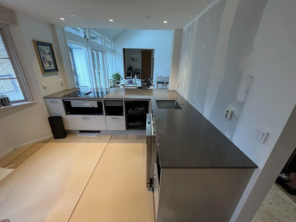 U-formad köksbänkskiva i mörk polerad Dekton Korso med halvö och ljusa köksstommar under montering i Kummelnäs.