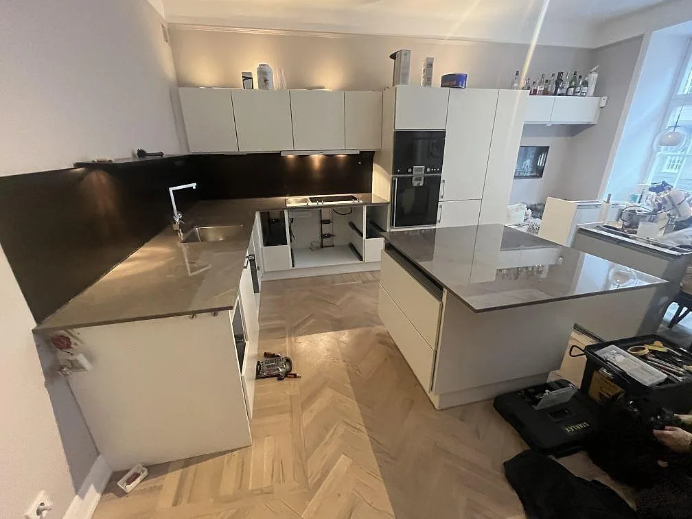 Köksbänkskiva i mörk polerad Dekton Korso med U-format bänk och köksö i ett kök med vita luckor i Stockholm under montering.