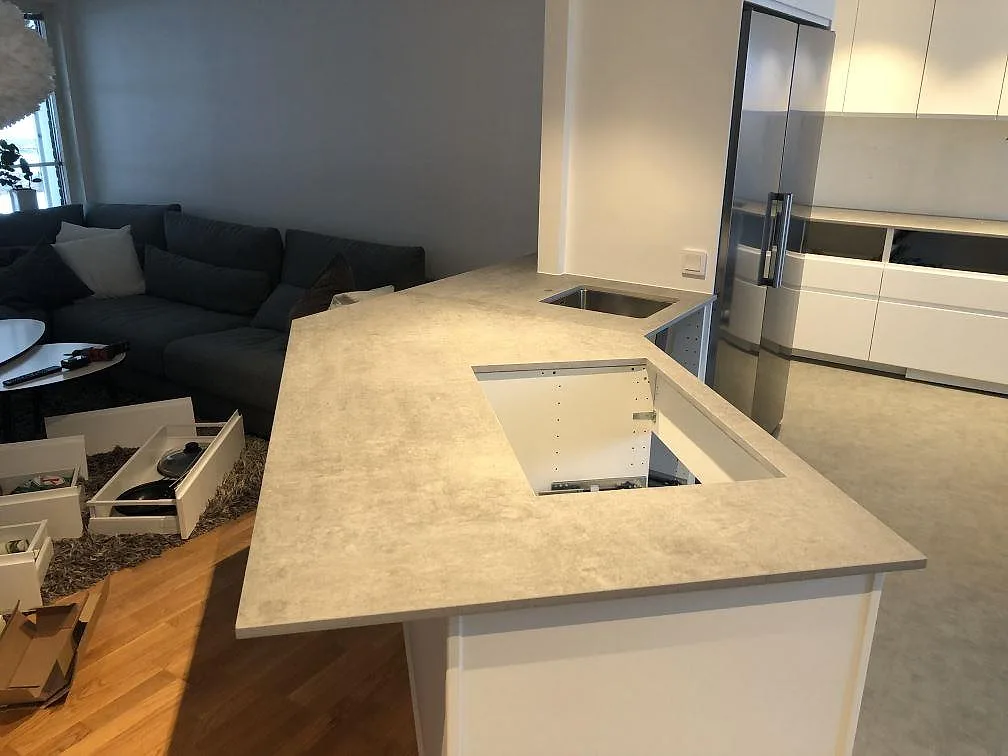 Köksbänkskiva i ljusgrå Dekton Kreta, honad matt yta, över en halvö med förberedd plats för häll och underlimmad rostfri diskho i ett kök i Hörby.