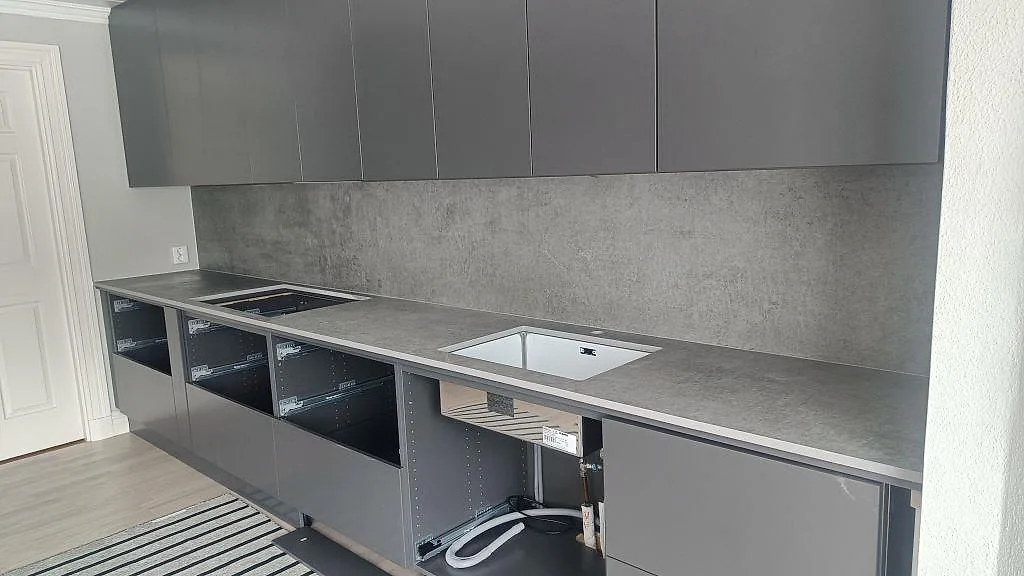 Köksbänkskiva i grå Dekton Kreta, matt betonglook, med heltäckande stänkskydd, underlimmad diskho och urtag för induktionshäll i ett kök under montering i Mullsjö.