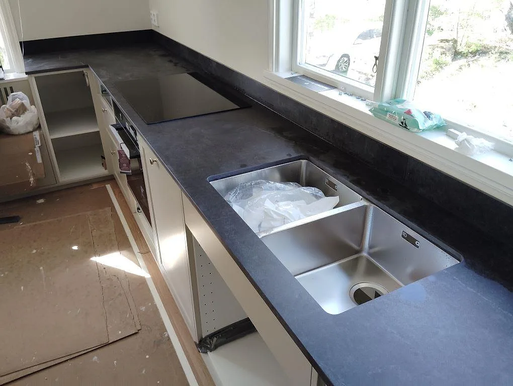 Köksbänkskiva i mörk matt Dekton Laos med underlimmad dubbel diskho, häll och stänkskydd vid ett fönster i Sigtuna.
