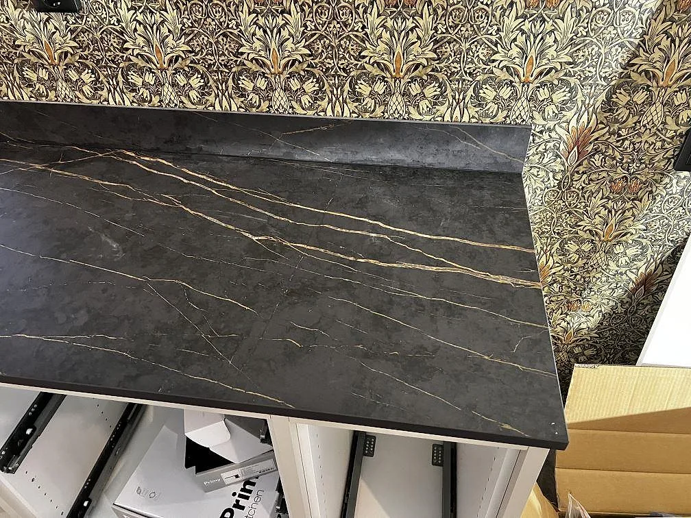 Närbild på matt Dekton Laurent keramik med svart botten, guldådring och uppvikt stänkkant mot blommig tapet i Gränna.