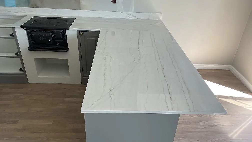 Polerad köksbänkskiva i Dekton Limbo med ljus ådring över halvön och svart spiszon i ett kök i Örby.