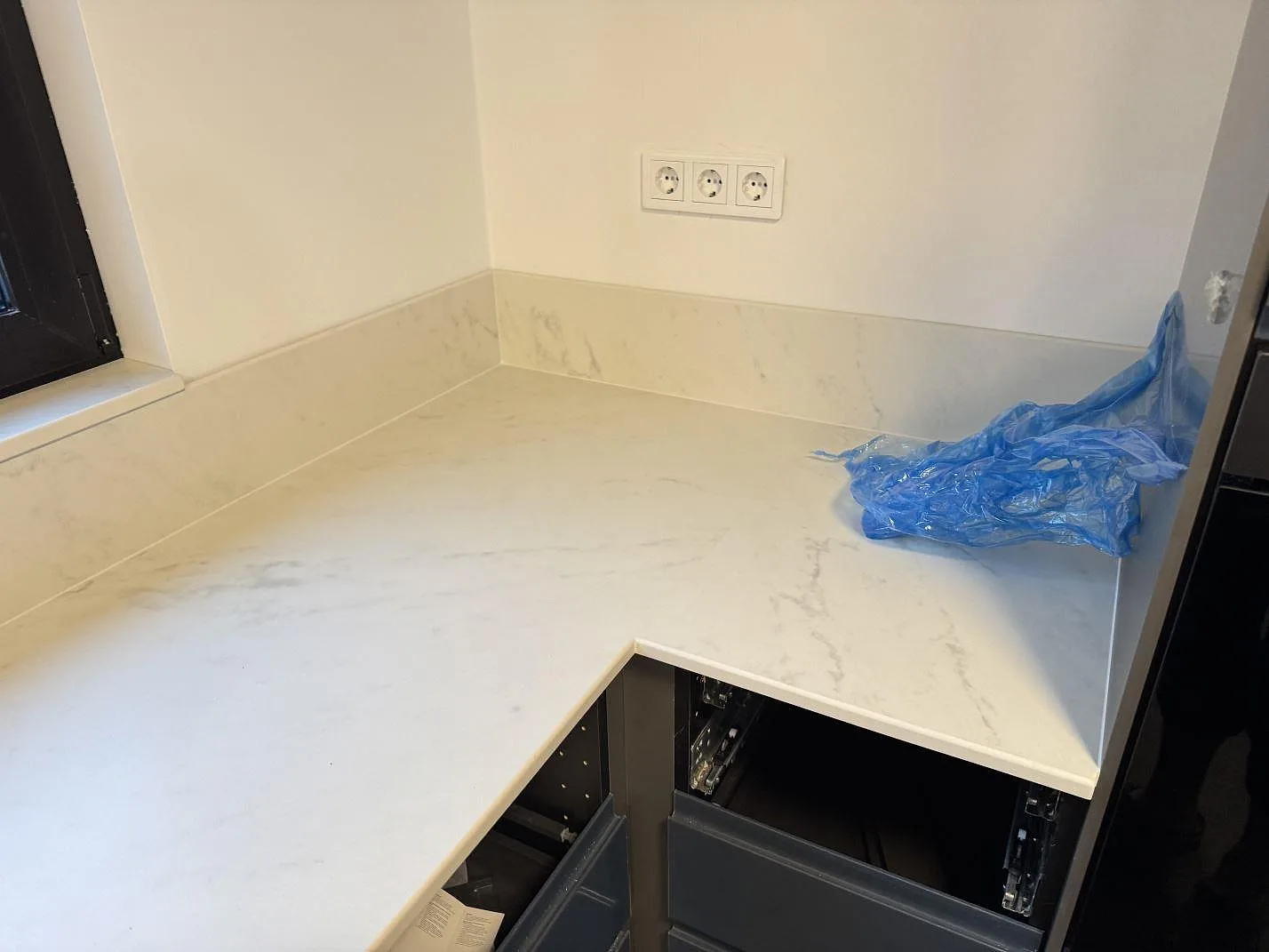 L-formad köksbänkskiva i ljus Dekton Marina Kc Velvet, matt, med stänkskydd och öppna köksstommar under montering i Lund.