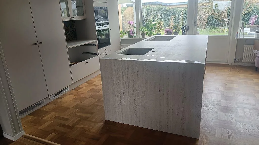 Köksö med bänkskiva i matt Dekton Marmorio i ett ljust kök i Limhamn, med diskho och hällöppning i den vitgrå stenen.