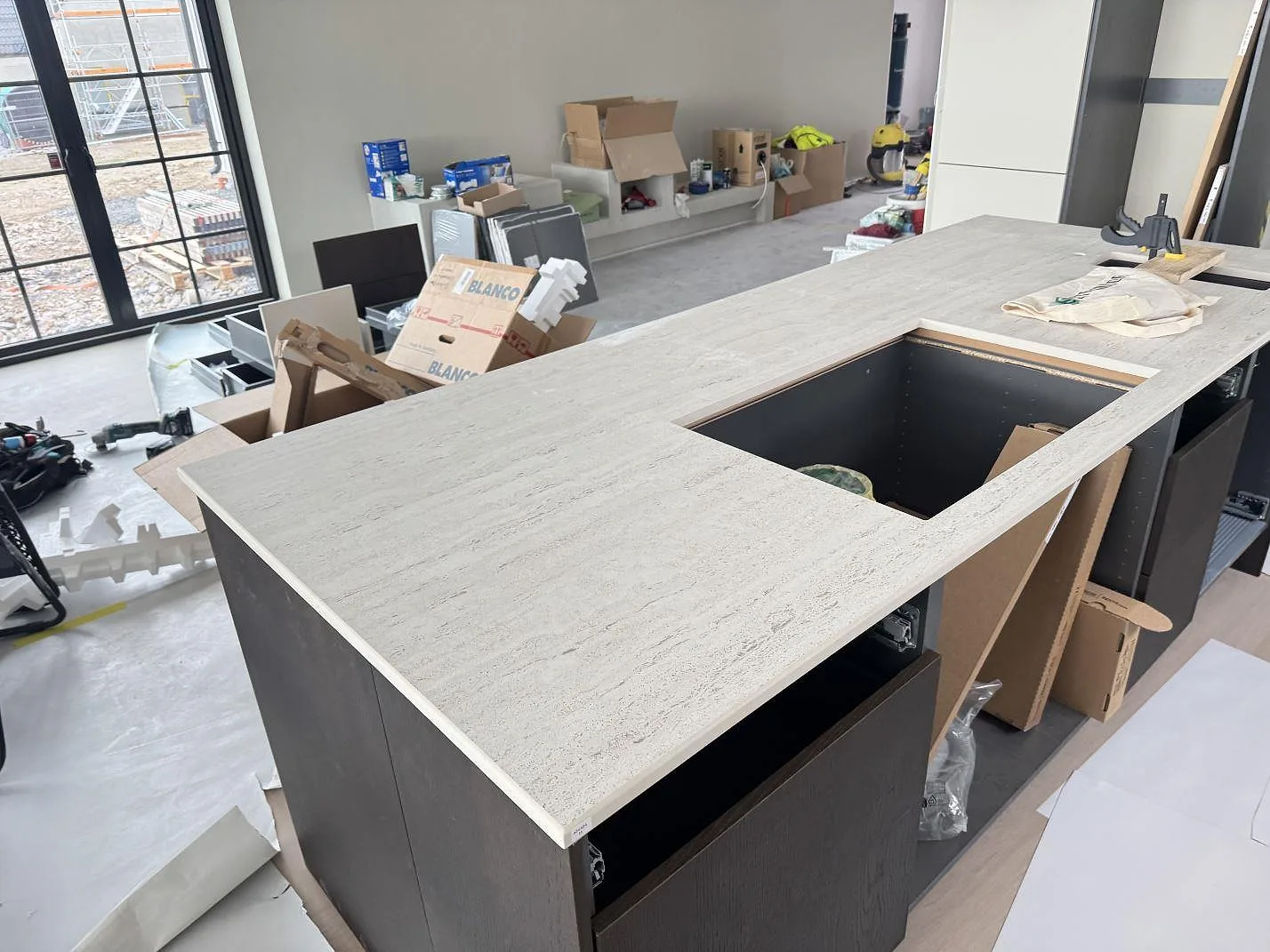 Bred köksö i matt Dekton Marmorio keramik med ljus linjär ådring och stort förberett urtag i ett kök under montering i Skurup.