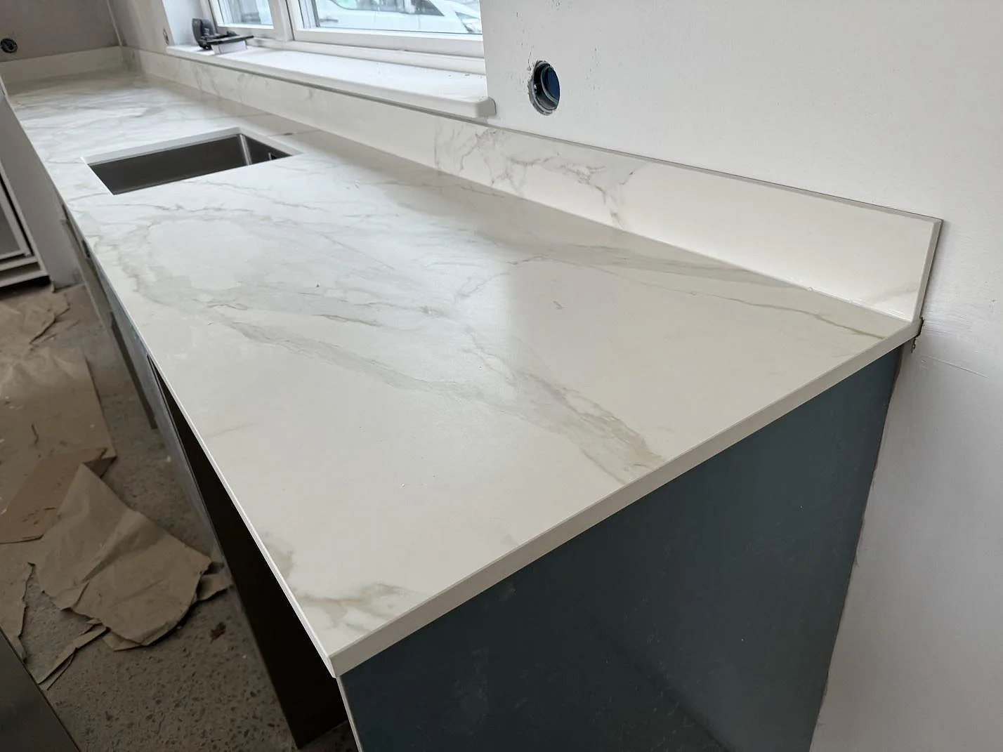 Lång köksbänkskiva i matt Dekton Morpheus Velvet med marmorerad ådring, låg stänkkant och fönsterparti under montering i Billesholm.