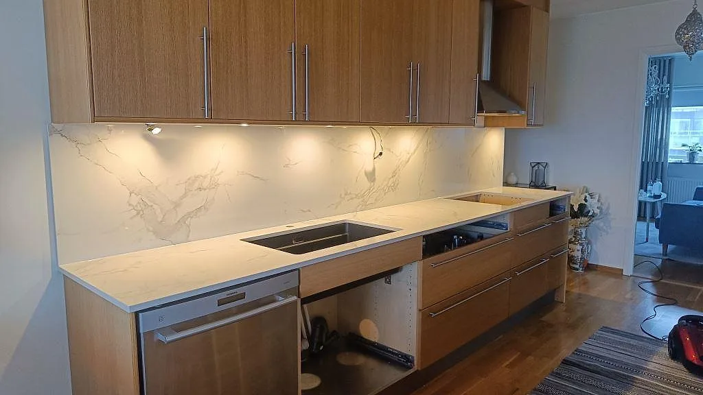 Köksbänkskiva i polerad Dekton Natura med hög marmorerad stänkvägg, underlimmad diskho och hällurtag i ett träkök i Limhamn.