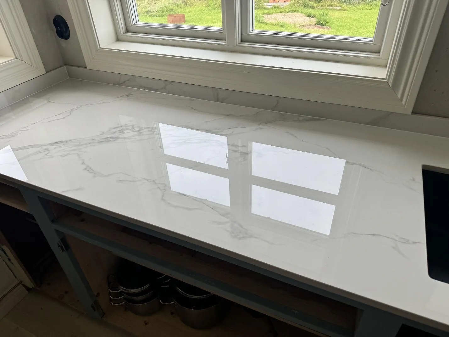 Lång köksbänkskiva i polerad Dekton Natura med grå ådring och låg stänkkant vid fönster i Tjureda.