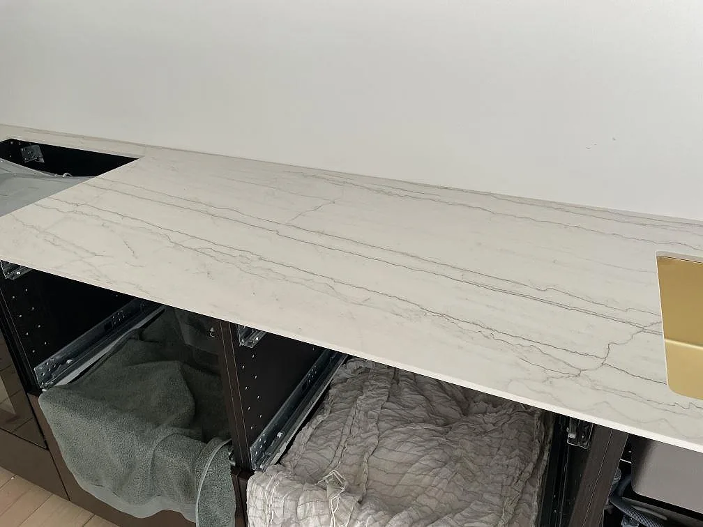 Köksbänkskiva i ljus matt Dekton Neural med grå ådring, öppen stomme och guldtonad diskho i Frederiksberg.