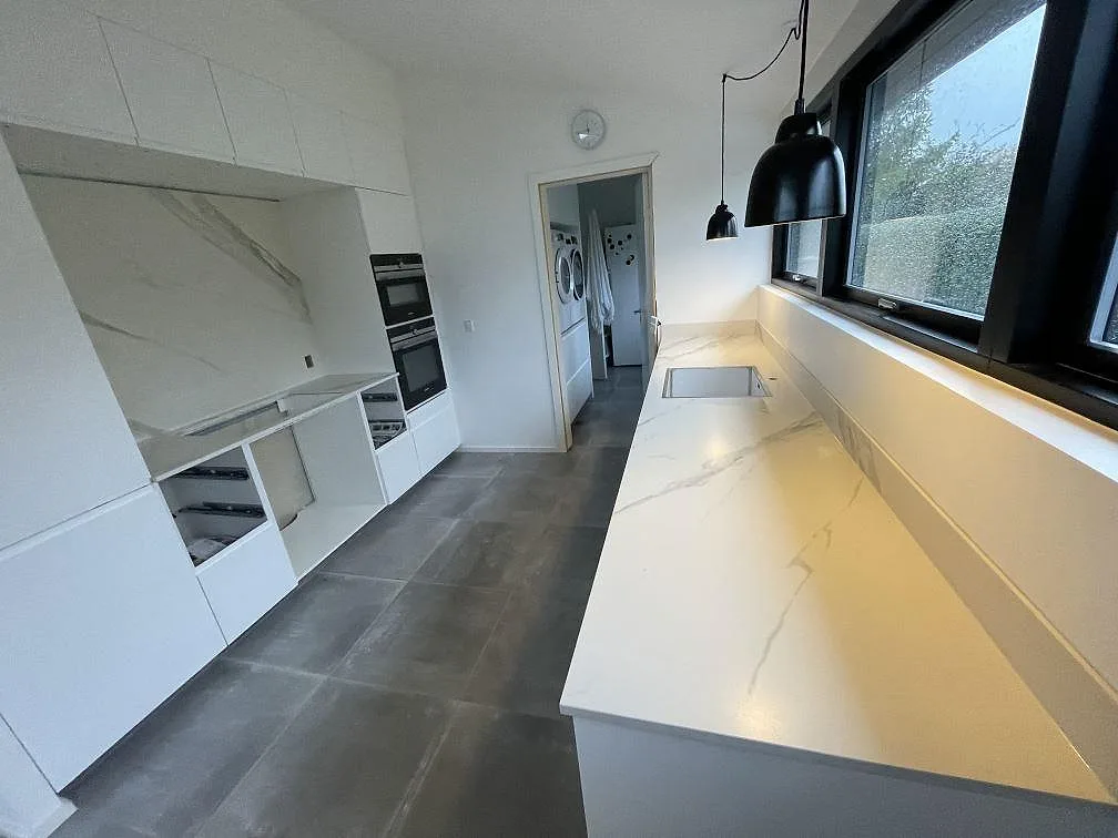 L-format köksbänkskiva i vit matt Dekton Opera Velvet keramik med rostfri underlimmad diskho, svarta fönster och mörka pendlar i Solroed Strand.