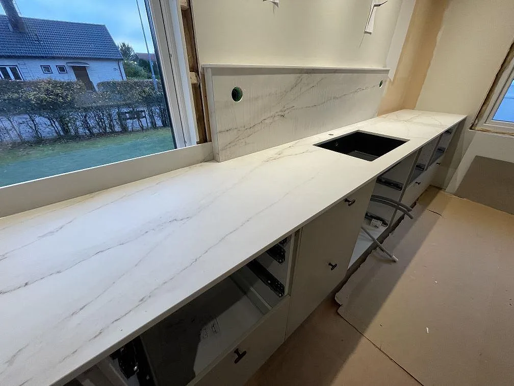 Lång köksbänk i matt Dekton Rem (Velvet) med svart diskho, blandarhål och uppvikt stänkskydd vid fönster i Kristinehamn.