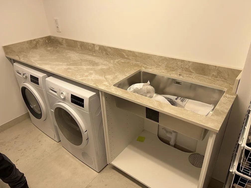 Krämvit Dekton Sarey i matt yta som arbetsbänk i en tvättstuga, med underlimmad rostfri diskho och plats för tvättmaskiner, i Bromma.