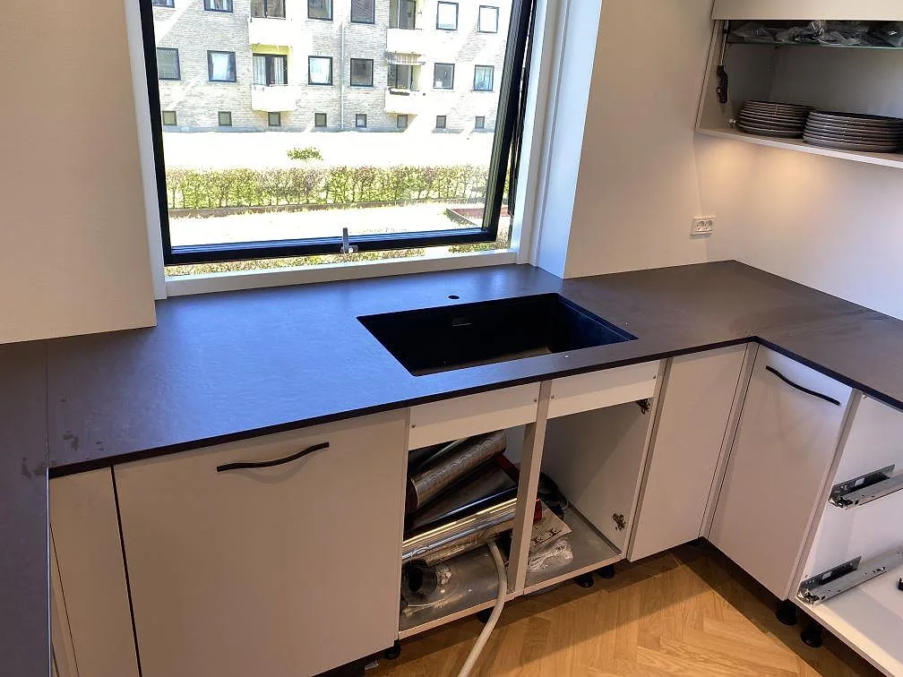 Matt köksbänkskiva i Dekton Sirius med svart diskho under fönstret, öppna köksstommar och mörk keramisk yta i Brønshøj.