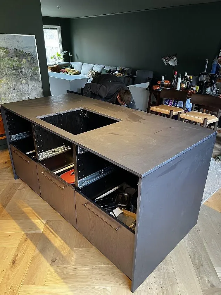 Köksö med mörk matt Dekton Sirius som keramikbänkskiva, med urtag i stenen och träinredning i ett kök under montering i Karlshamn.