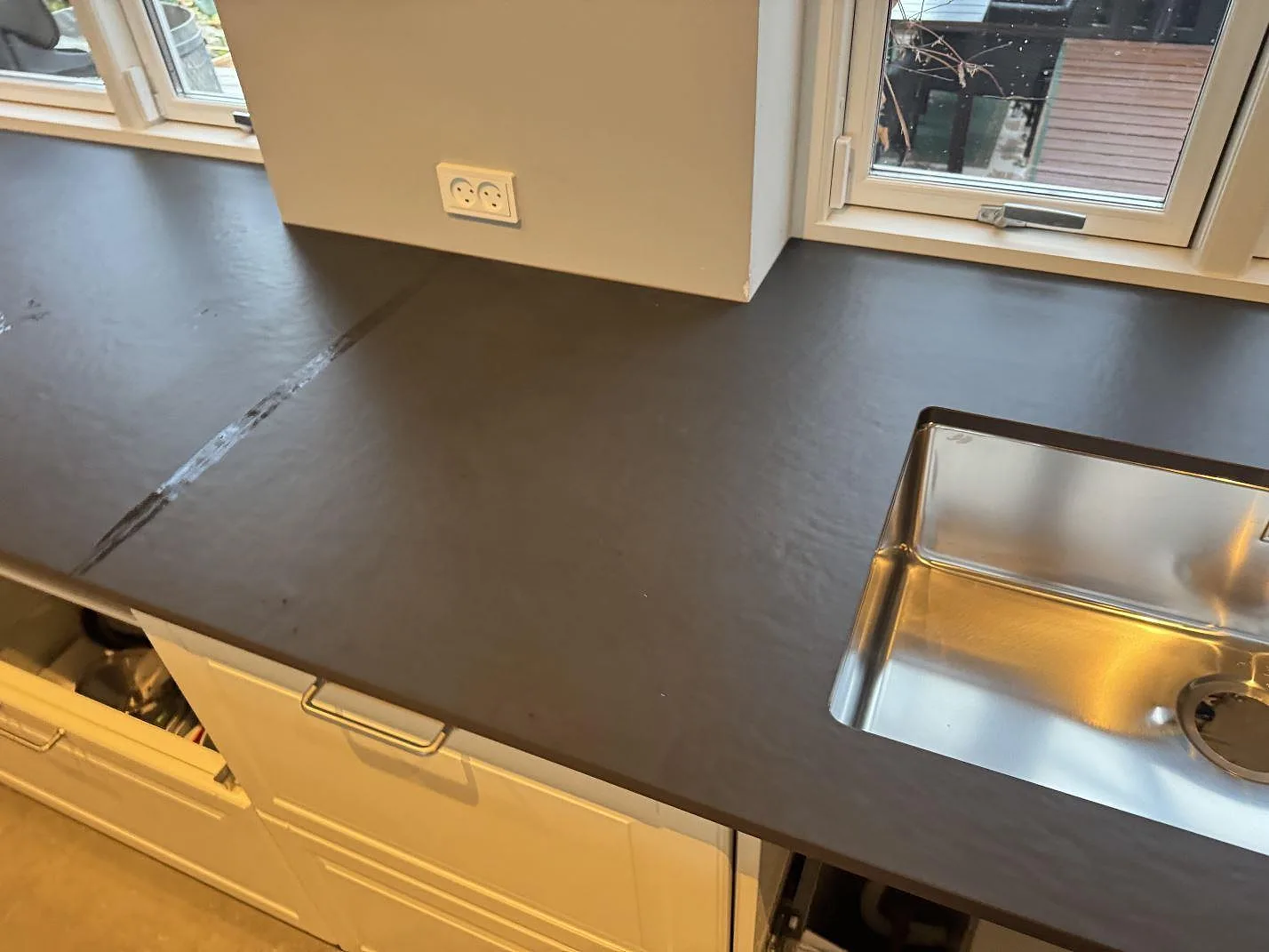 Köksbänkskiva i mörkt grafitgrå Dekton Sirius, matt yta, med stor underlimmad rostfri diskho och köksfönster mot trädgården i ett kök under montering i Mørkøv.