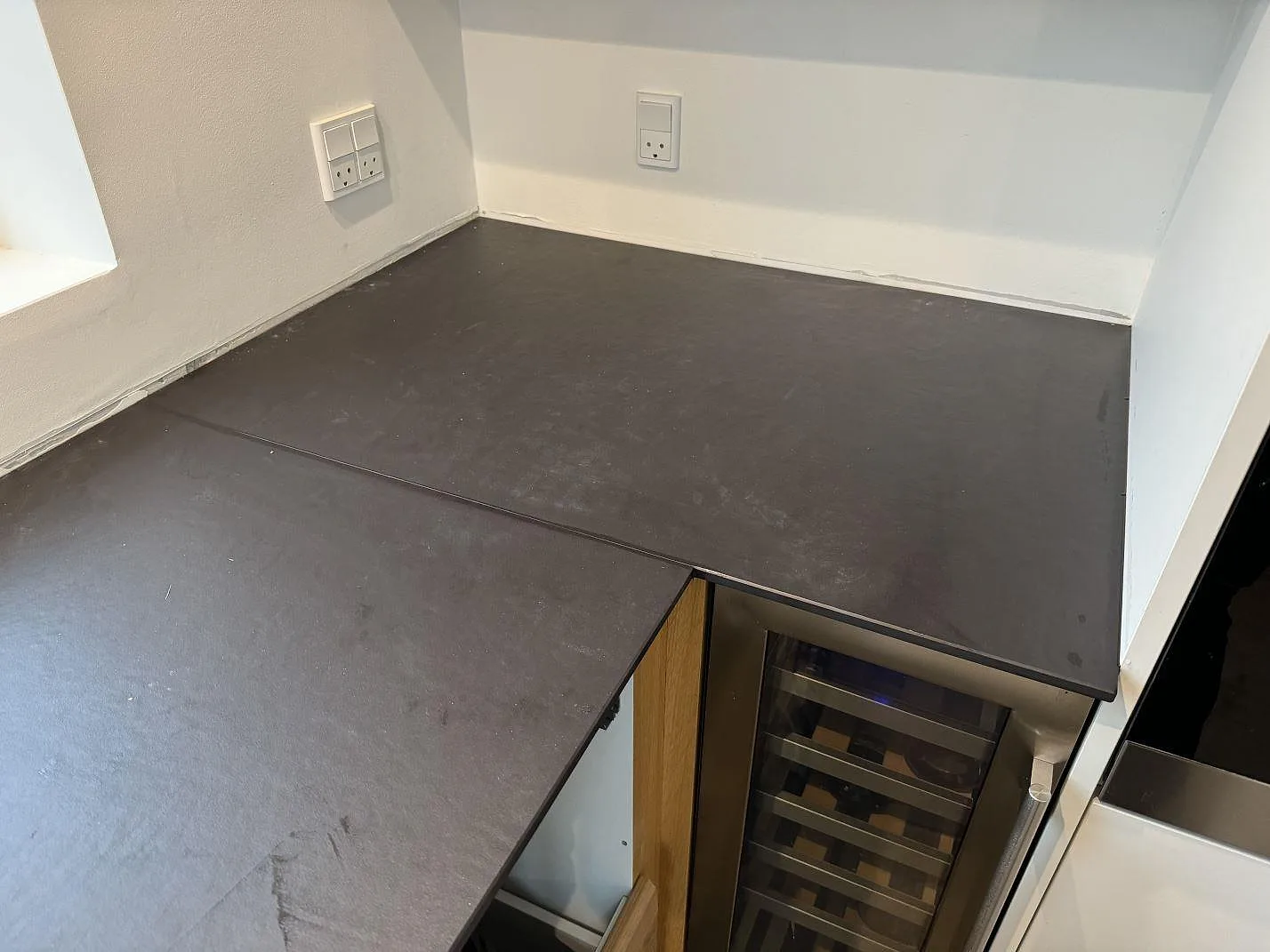 L-formad köksbänkskiva i mörk matt Dekton Sirius under fönster och vägguttag i Roskilde.