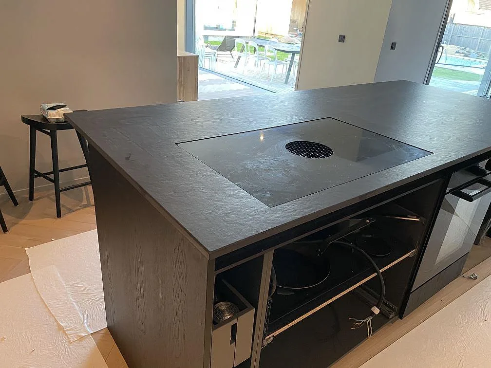 Köksbänkskiva och köksö i mörk Dekton Sirius, matt, med nedfälld induktionshäll och integrerad fläkt i ett kök under montering i Särö.