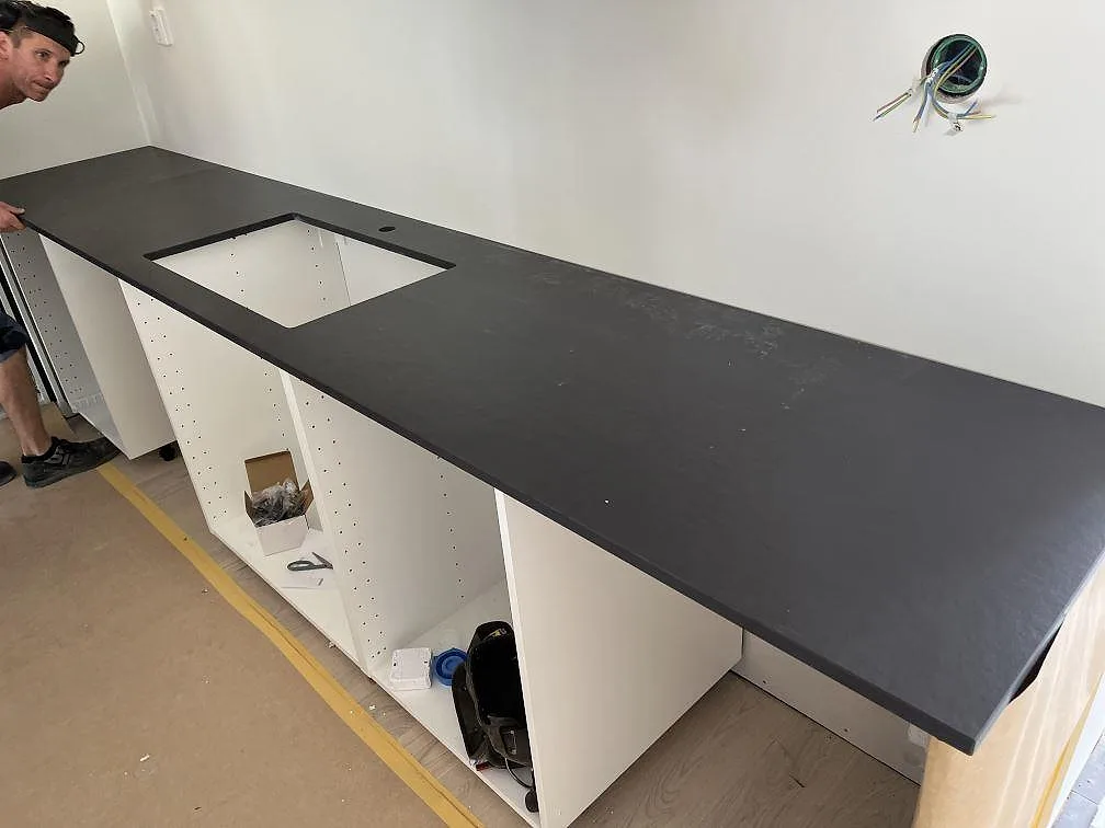 Köksbänkskiva i mörk Dekton Sirius, matt keramik, monterad på vita köksstommar med urtag för underlimmad diskho i ett kök under installation i Växjö.