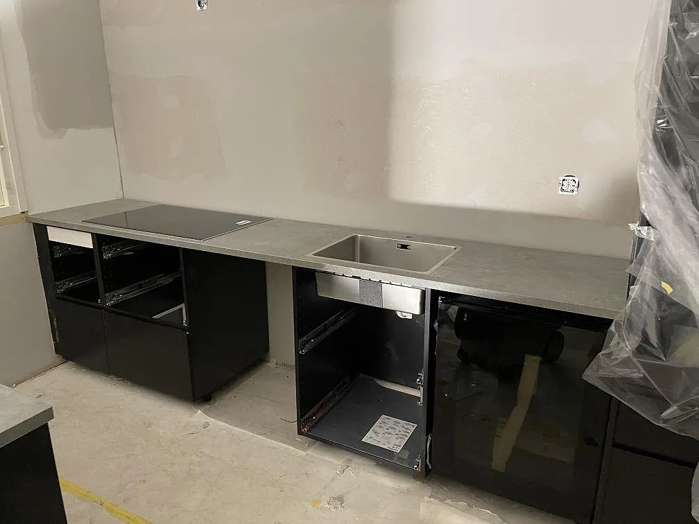 Köksbänkskiva i grå Dekton Soke, matt betonglook, med övermonterad induktionshäll och planmonterad rostfri diskho i ett kök under montering i Arrie.