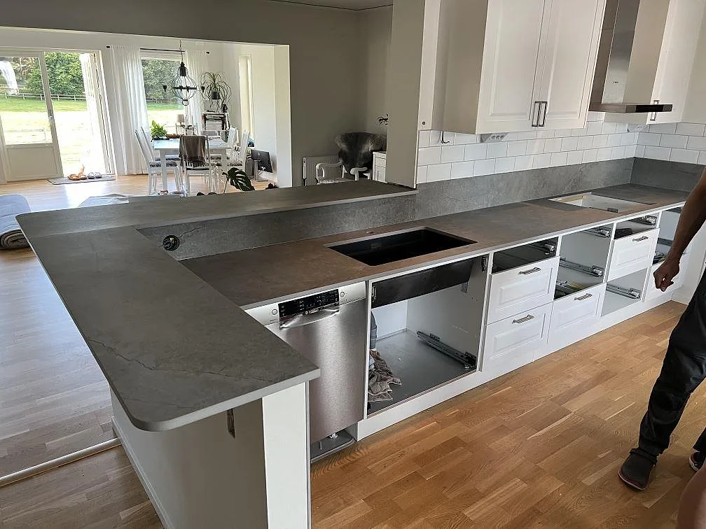 Köksbänkskiva i matt grå Dekton Soke, keramik, med stänkskydd och öppna köksstommar i ett kök under montering i Flyinge.