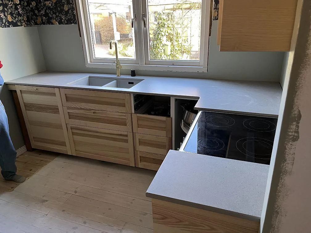 Ljusgrå köksbänkskiva i keramik (Dekton Strato) med matt yta monterad som rak köksrad längs fönstret i ett kök i Lund, med underlimmad dubbelho och borstad mässingsblandare under montering.