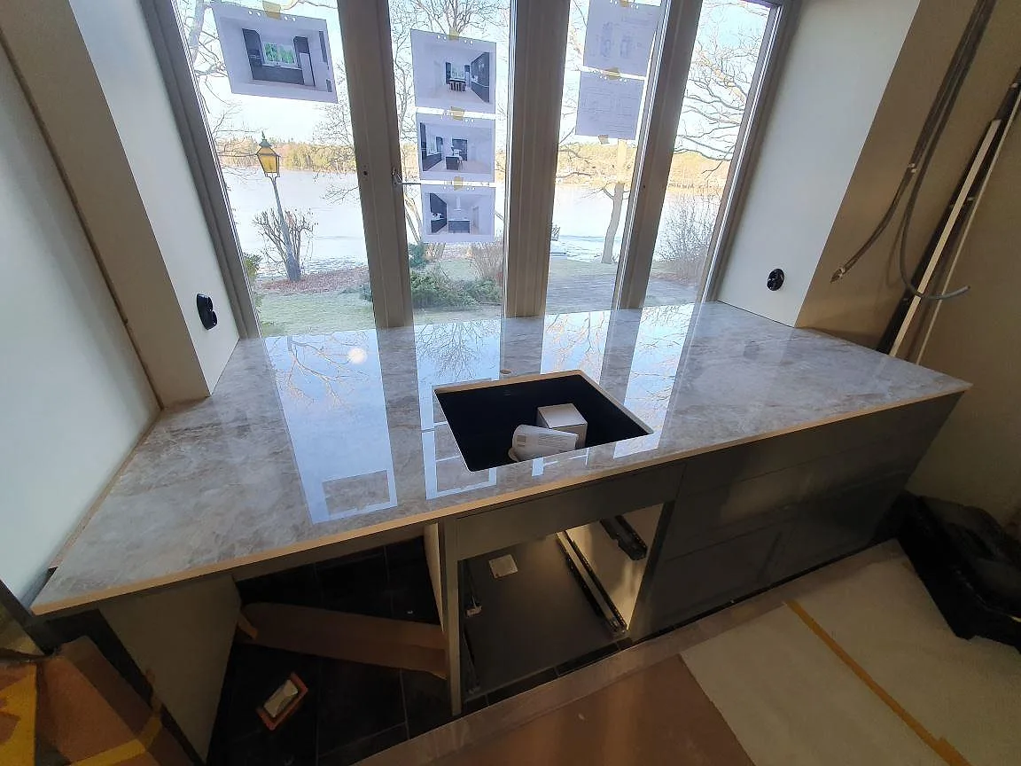 Polerad köksbänkskiva i Dekton Taga, varm greige keramik med underlimmad diskho, vid ett fönsterparti med sjöutsikt i ett kök i Älta.
