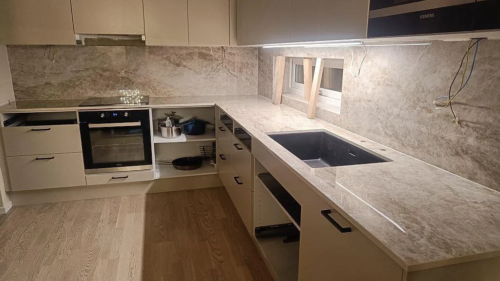 Köksbänkskiva i varmgrå Dekton Taga, polerad, i ett L-format kök med heltäckande stänkvägg, underlimmad svart komposithon och infälld induktionshäll i Borås.