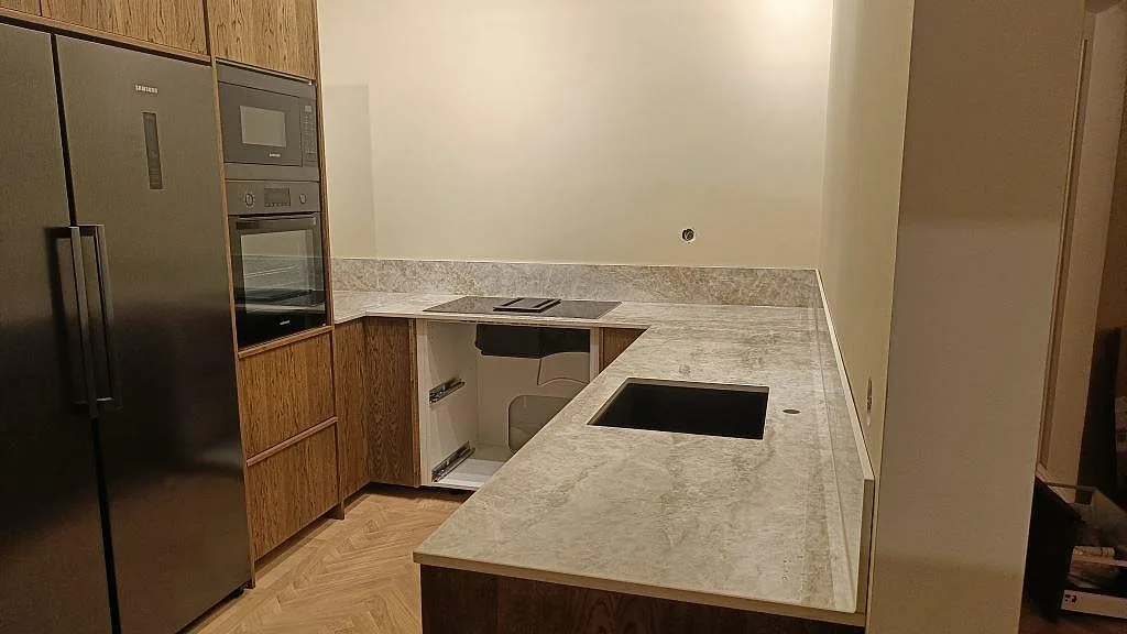 Köksbänkskiva i Dekton Taga, ljusgrå polerad keramik, i ett U-format kök i Göteborg med underlimmad svart diskho, svart induktionshäll och ekluckor.