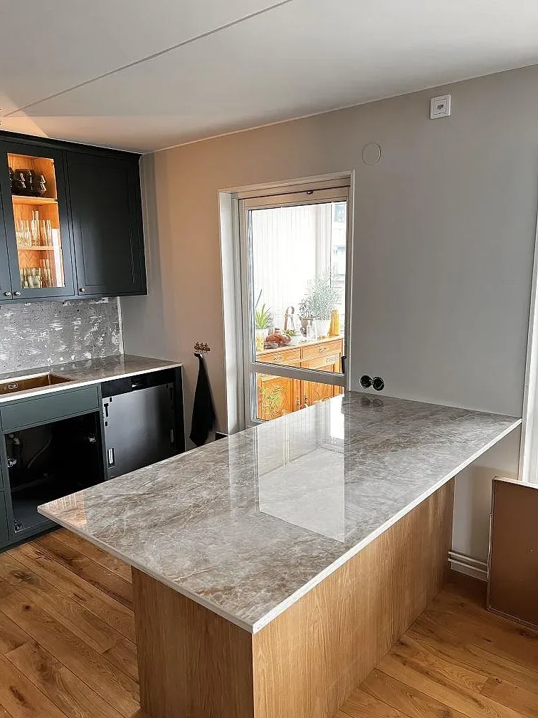 Köksö med polerad Dekton Taga keramik i gråbeige marmorlook, träinredning och mörkgröna stommar i Limhamn.