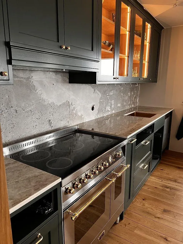 Köksbänkskiva i polerad Dekton Taga keramik med stänkskydd vid svart spis i ett kök med mörkgröna luckor i Limhamn.