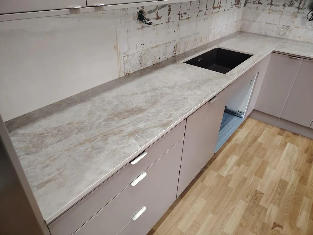 Närbild av polerad Dekton Taga köksbänkskiva med svart diskho, ljus marmorering och öppna köksstommar under montering i Norsborg.