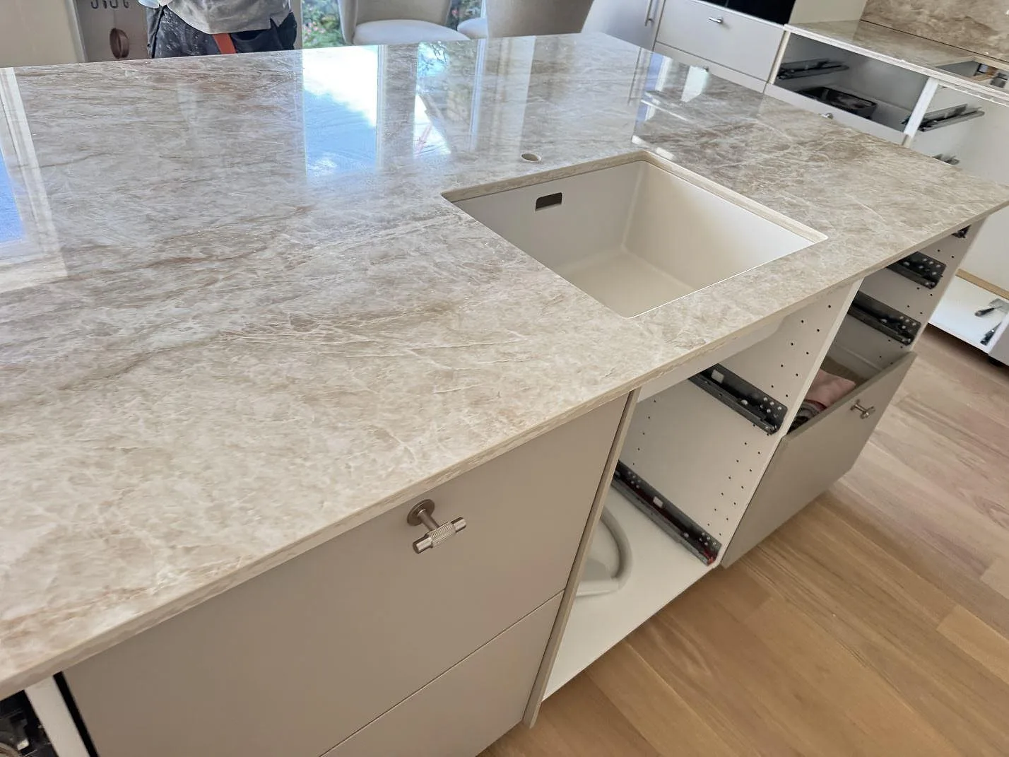 Närbild på vit diskho i en polerad Dekton Taga köksbänkskiva med ljus ådring och öppna köksstommar i Virum.