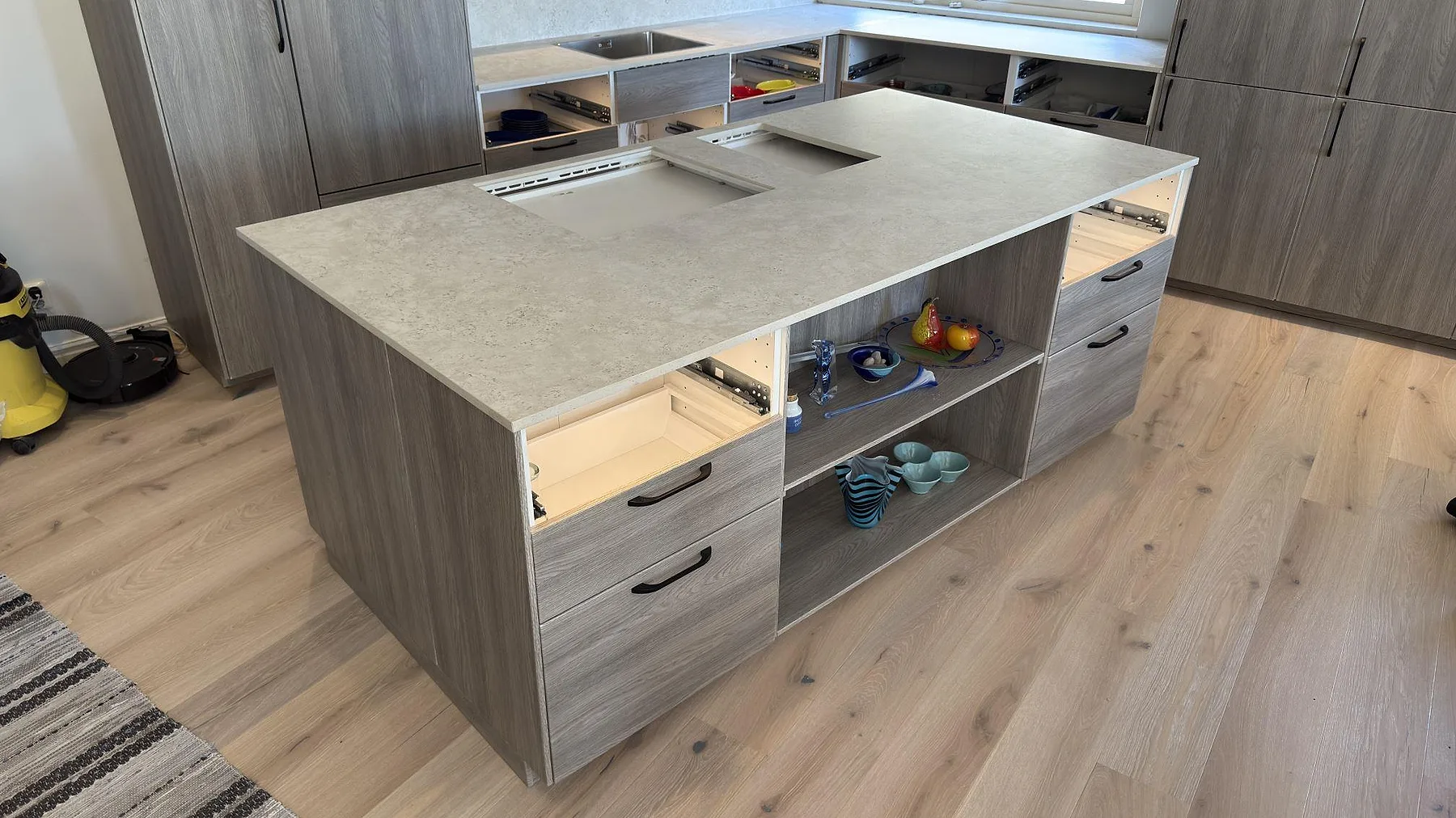 Stor fristående köksö i matt Dekton TK05 Sabbia med öppna lådstommar och plats för häll i ett kök under montering i Åkersberga.