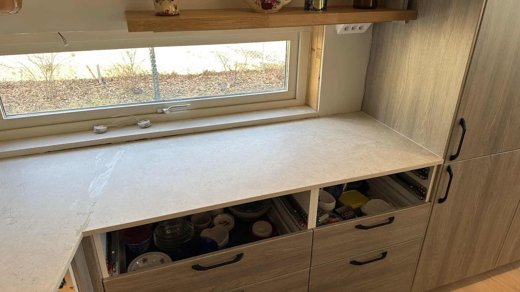 Köksbänkskiva i matt Dekton TK05 Sabbia under ett fönster, sandgrå keramik med ljusa träfronter i ett kök i Åkersberga.