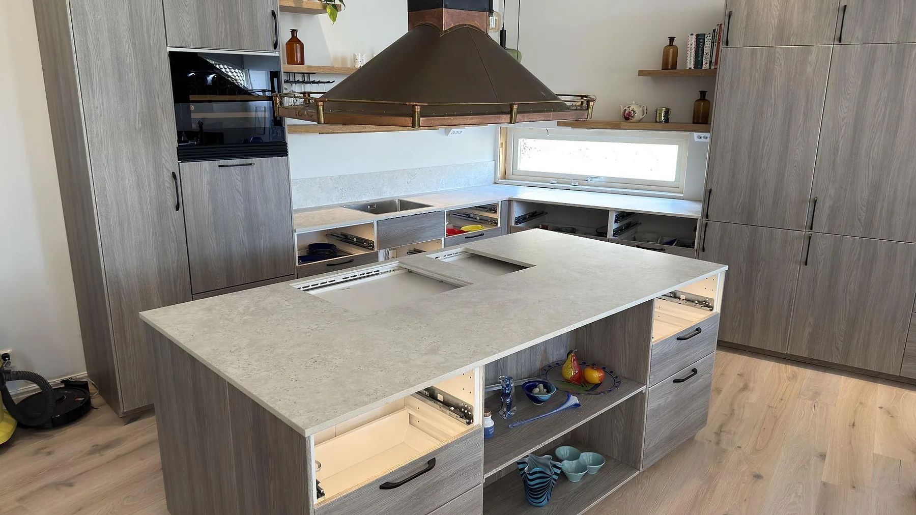 Köksö med köksbänkskiva i matt Dekton TK05 Sabbia, sandgrå keramik med urtagning för häll, under en kopparfärgad köksfläkt i ett kök i Åkersberga.
