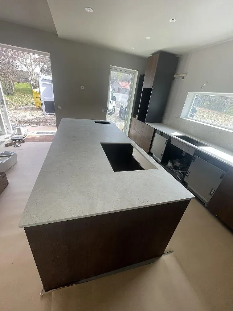 Köksö i ljus Dekton Tk05 Sabbia keramik med uppvikt stänkkant och valnötsfärgade luckor i ett kök i Alingsås.