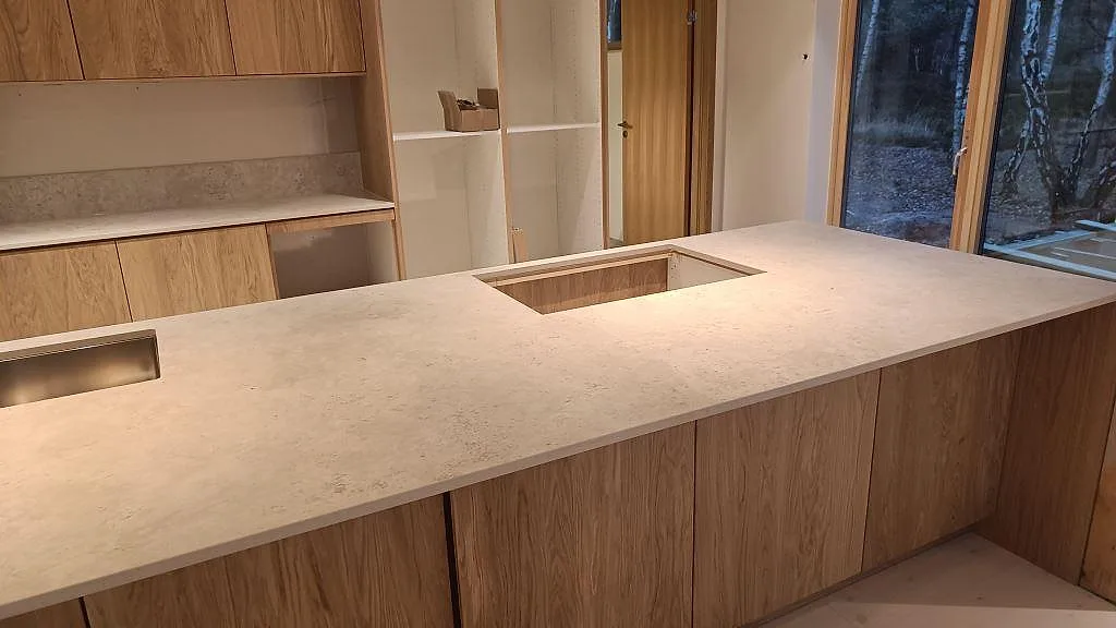 Lång köksö i matt Dekton Tk05 Sabbia med hällöppning och rostfri diskho i ett kök under montering i Höllviken.