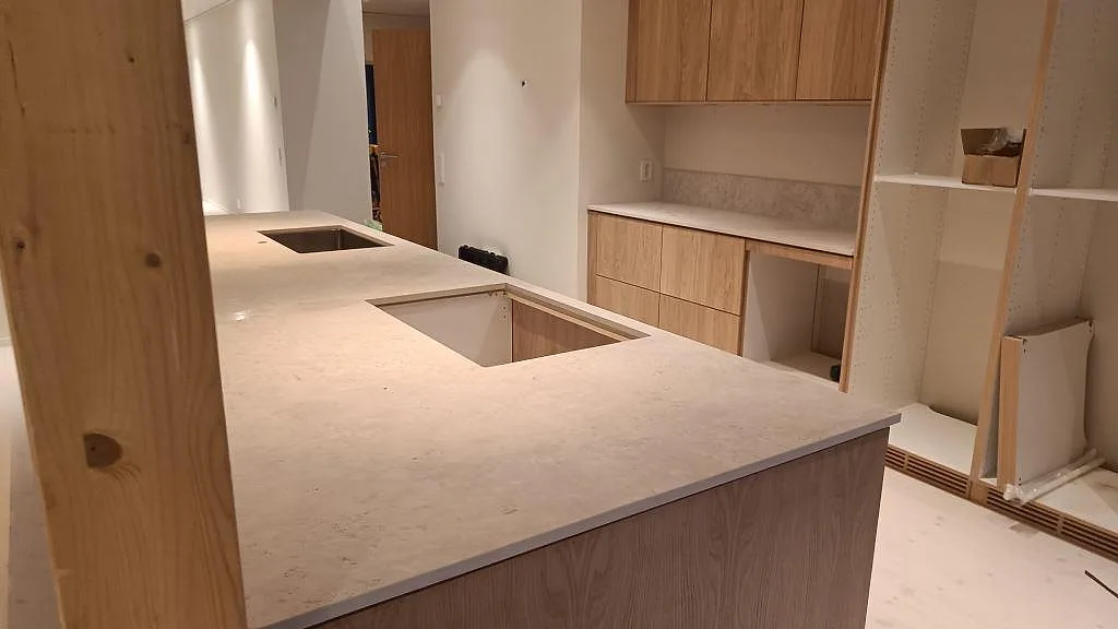 Köksö i matt Dekton Tk05 Sabbia med hällöppning, rostfri diskho och öppen inredning under montering i Höllviken.