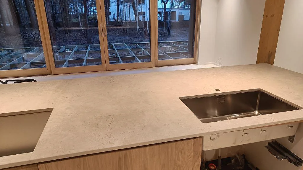 Bänkskiva i matt Dekton Tk05 Sabbia med rostfri diskho och hällöppning i köksö framför stora fönster i Höllviken.