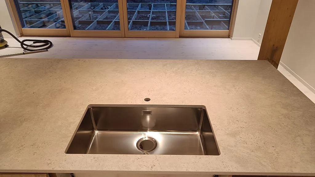 Rostfri diskho i en köksö med matt Dekton Tk05 Sabbia, beige keramisk yta och blandarhål i Höllviken.