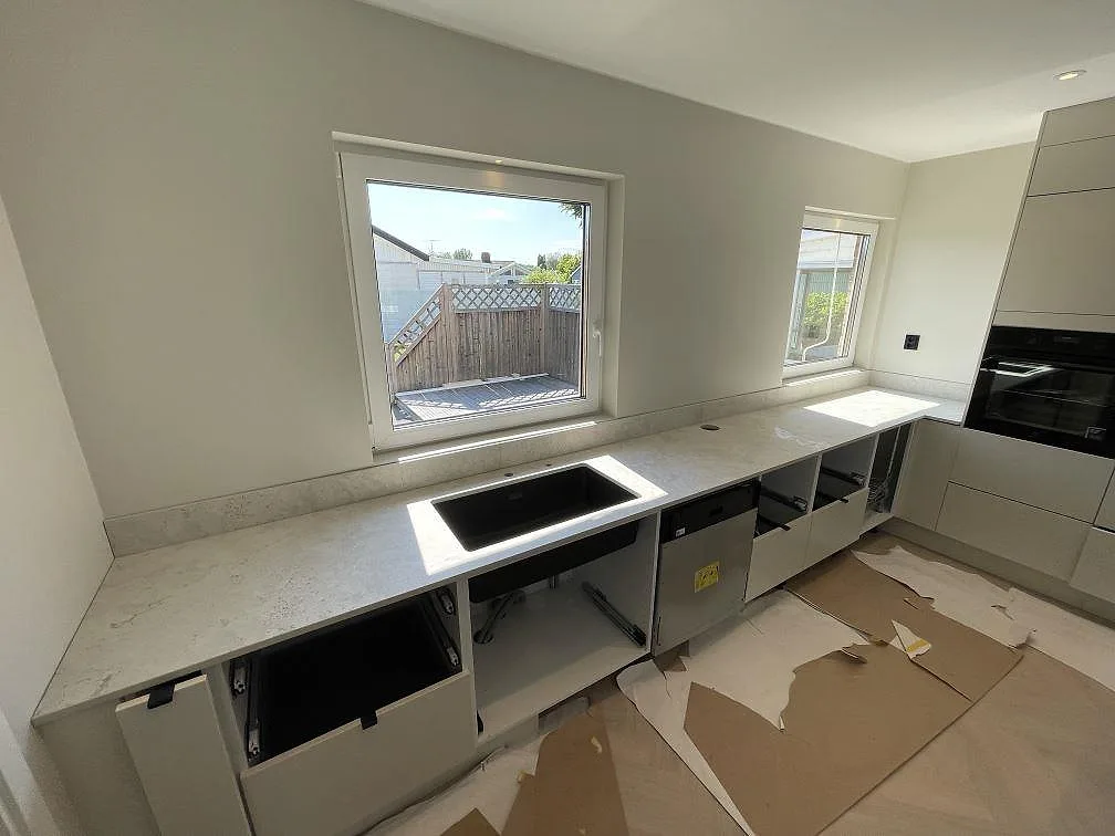 Lång köksbänkskiva i matt Dekton Tk05 Sabbia med svart diskho, stänkskydd och öppna köksstommar under montering i Örebro.