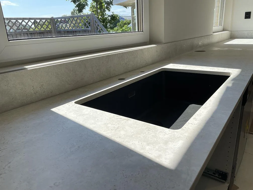 Närbild på svart diskho i köksbänkskiva av matt Dekton Tk05 Sabbia, med sandbeige keramikyta och stänkskydd vid fönster i Örebro.