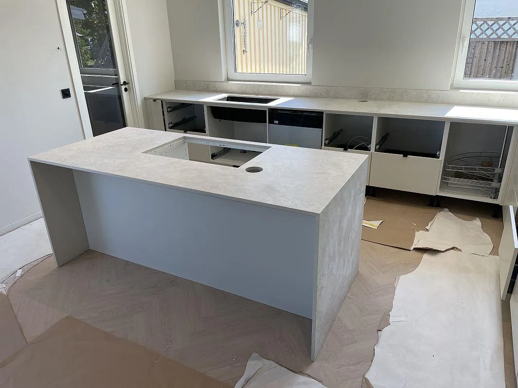 Bred vy över kök i Örebro med matt Dekton Tk05 Sabbia som köksö, bänkskiva under fönster och låg stänkkant i samma keramik.