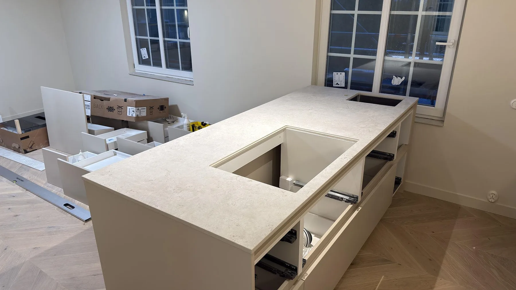 Köksö i matt Dekton Tk05 Sabbia med hällöppning och ljus sandtonad keramikyta i ett kök under montering i Stockholm.